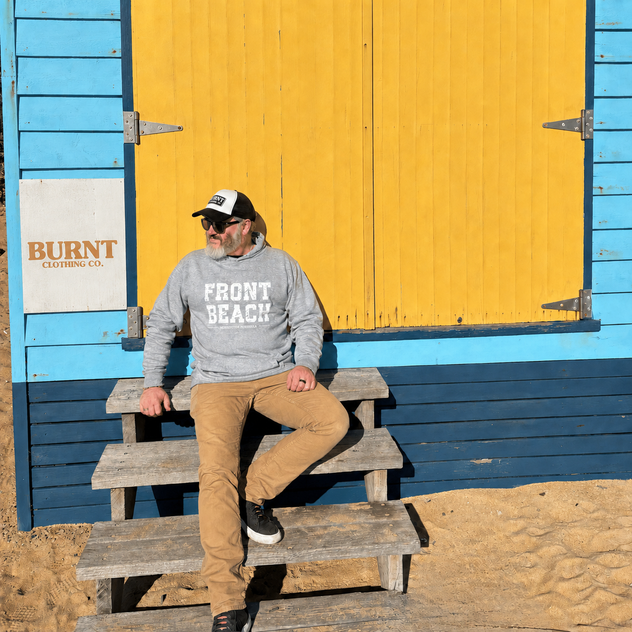 Man seated outside a colourful beach box wearing the Front Beach grey marle hoodie