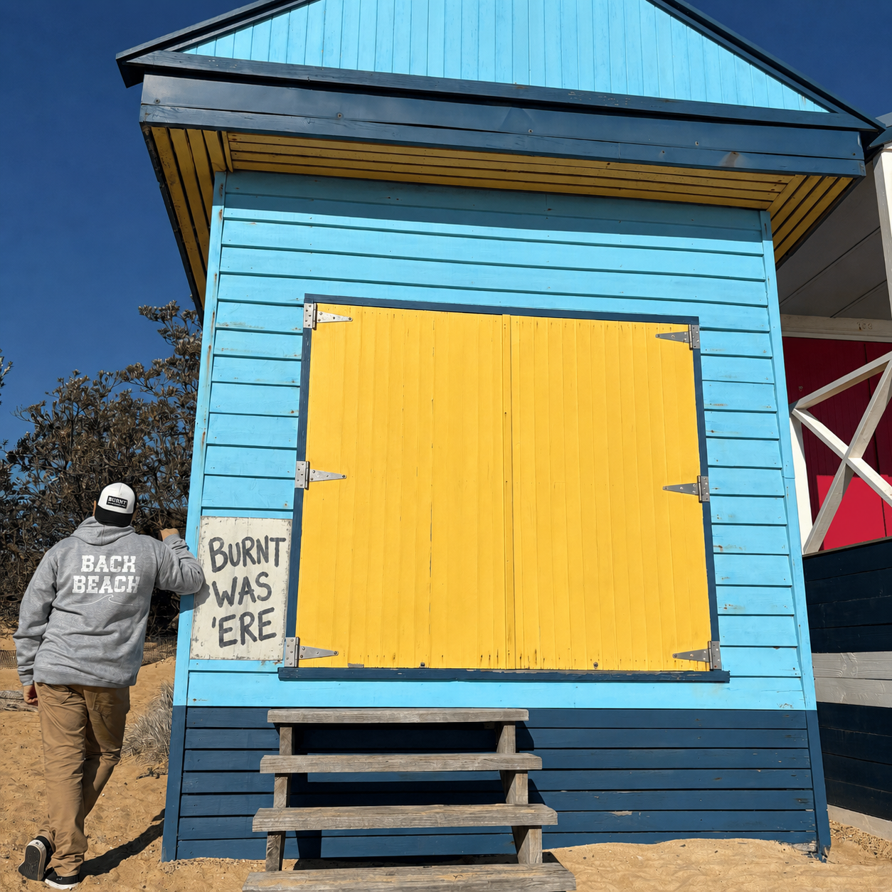 Man wearing the Back Beach grey marle hoodie beside a blue and yellow Mornington Peninsula beach box