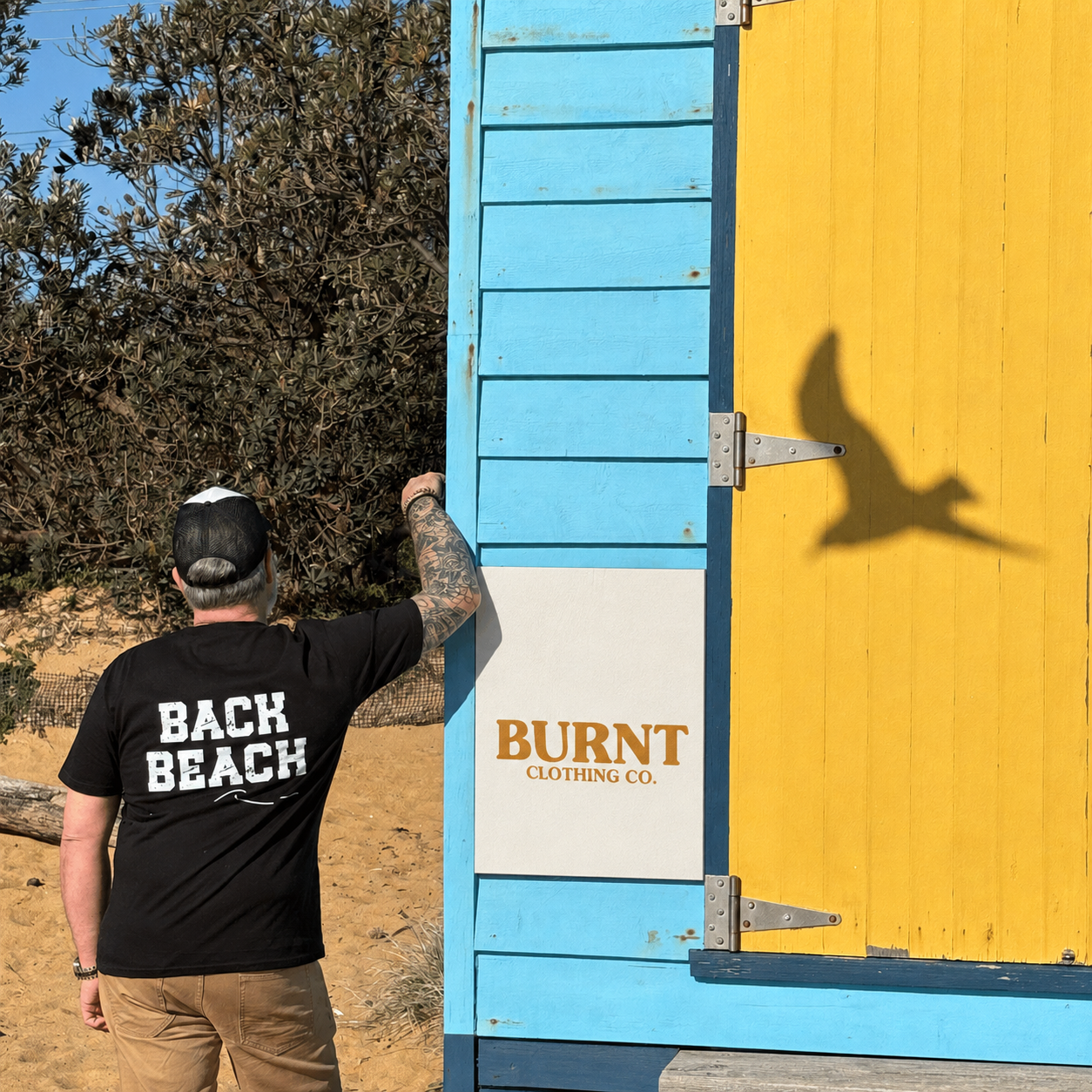 Rear view of the black tee showing the distressed Back Beach print and wave detail beside a colourful beach box