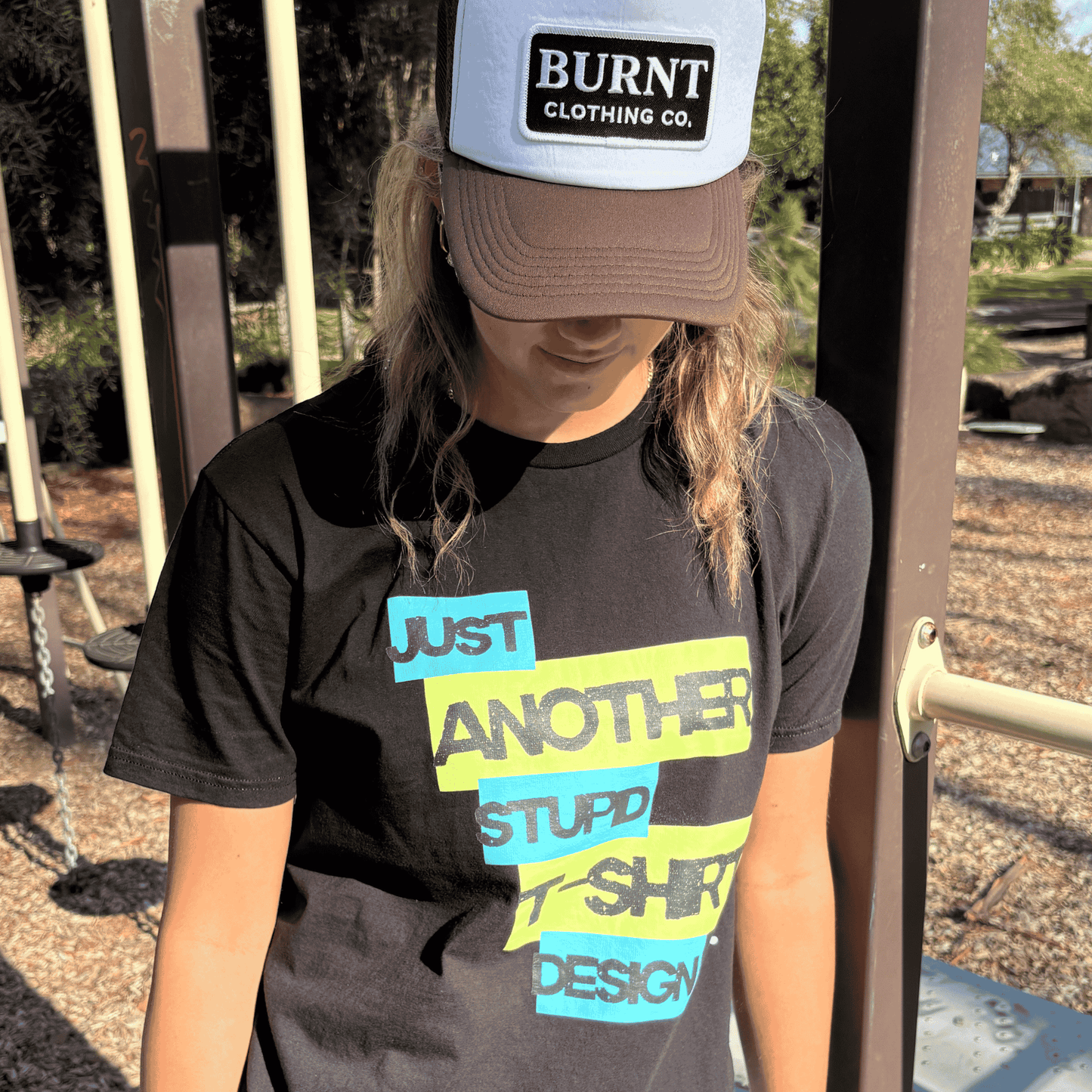Person wearing Burnt Just Another Stupid T-Shirt Design graphic tee in black with Burnt Clothing Co. Trucker Cap