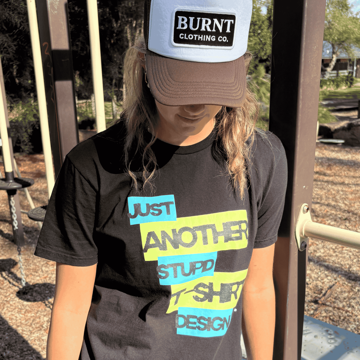 Person wearing Burnt Just Another Stupid T-Shirt Design graphic tee in black with Burnt Clothing Co. Trucker Cap