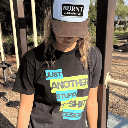 Person wearing Burnt Just Another Stupid T-Shirt Design graphic tee in black with Burnt Clothing Co. Trucker Cap