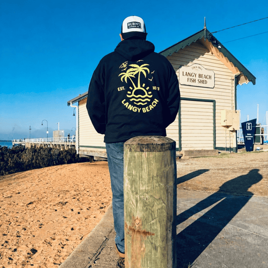 Person wearing a black hoodie with 'Langy Beach' branding, standing by a wooden post with a fish shed in the background.