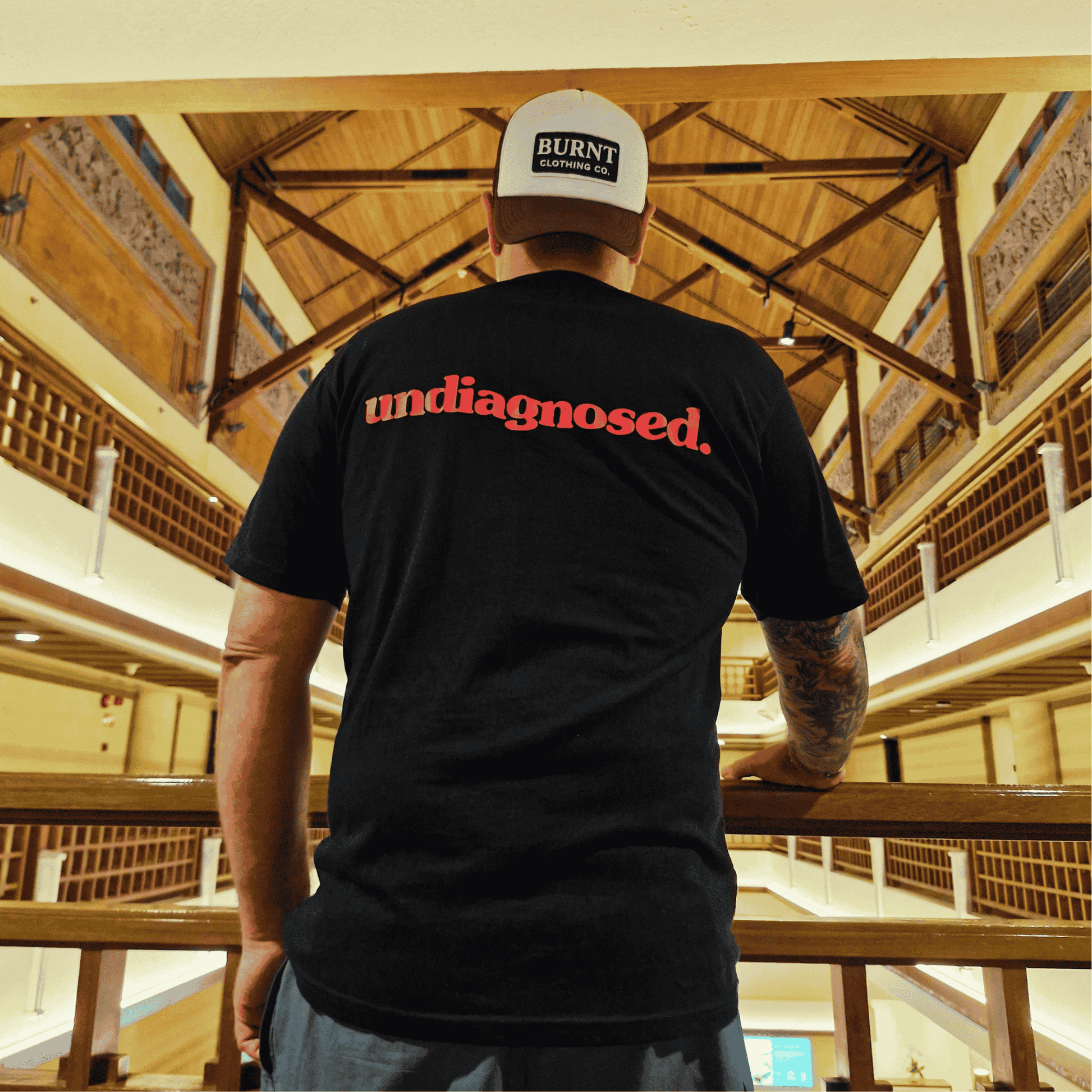 Person wearing a black t-shirt with 'undiagnosed' text and a white cap with 'Burnt' text, standing in a wooden architectural interior.