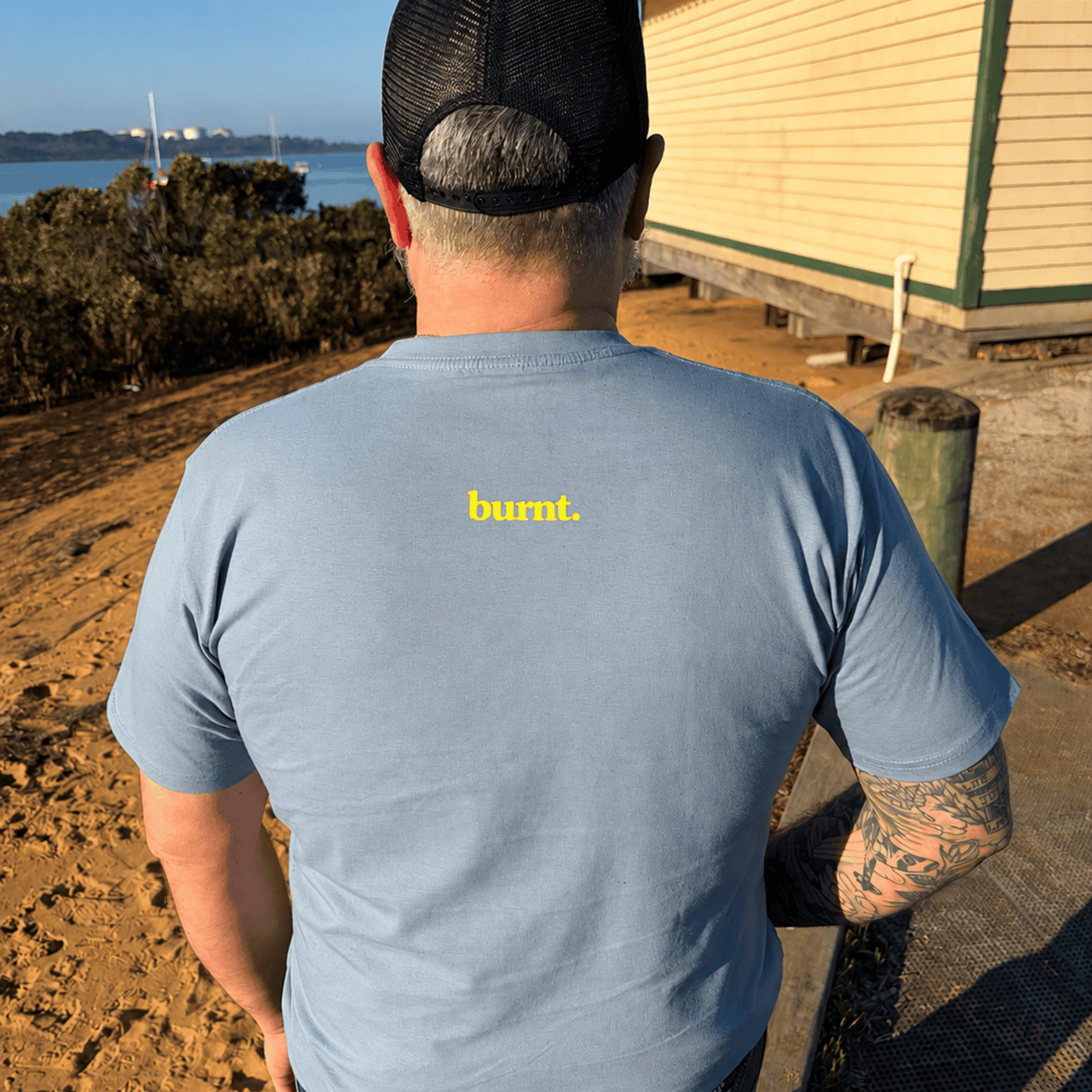 Back view of Langy Beach blue tee with yellow burnt. logo outdoors