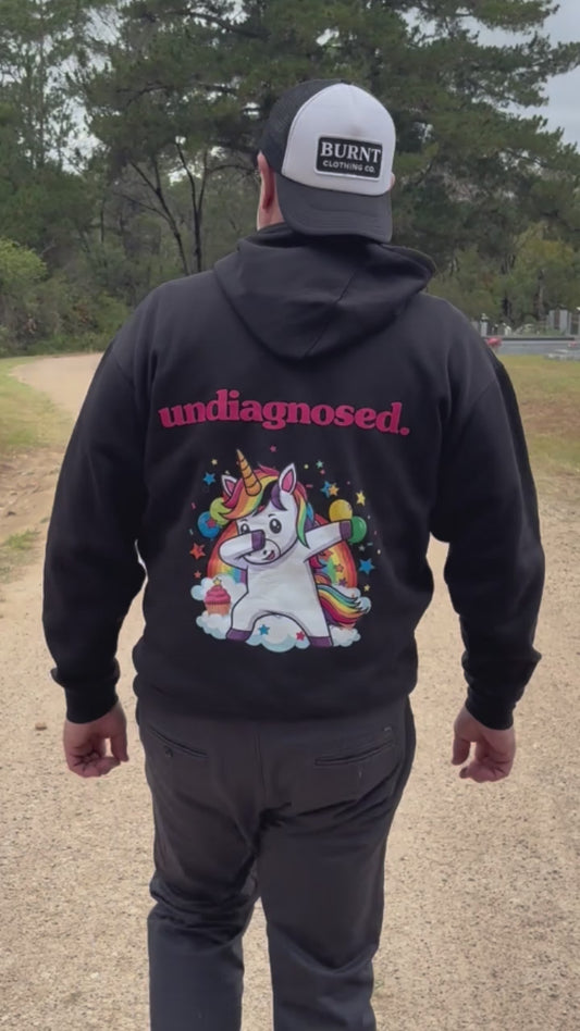 “Person walking away outdoors wearing a black undiagnosed. hoodie with pink lettering and a colourful unicorn graphic on the back.”