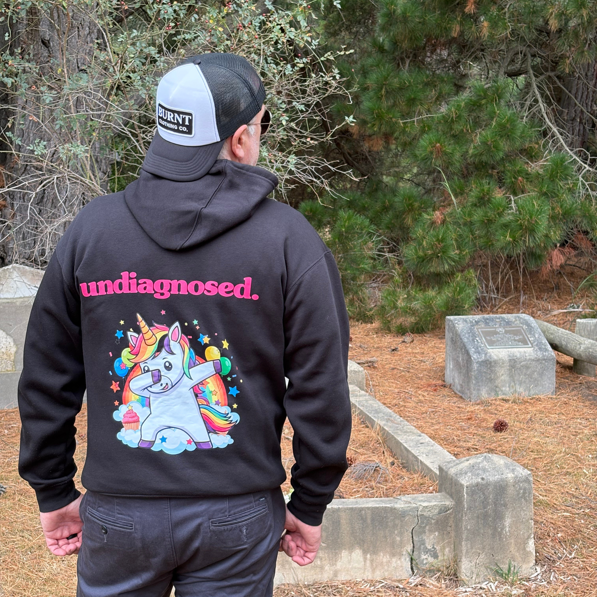 Person wearing a black undiagnosed. hoodie outdoors, showing pink lettering and a colourful unicorn graphic on the back.